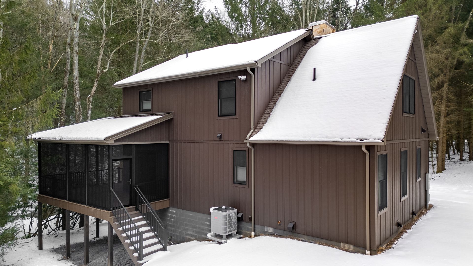 Snow-covered brown house with screened porch and steep roof in a wooded yard