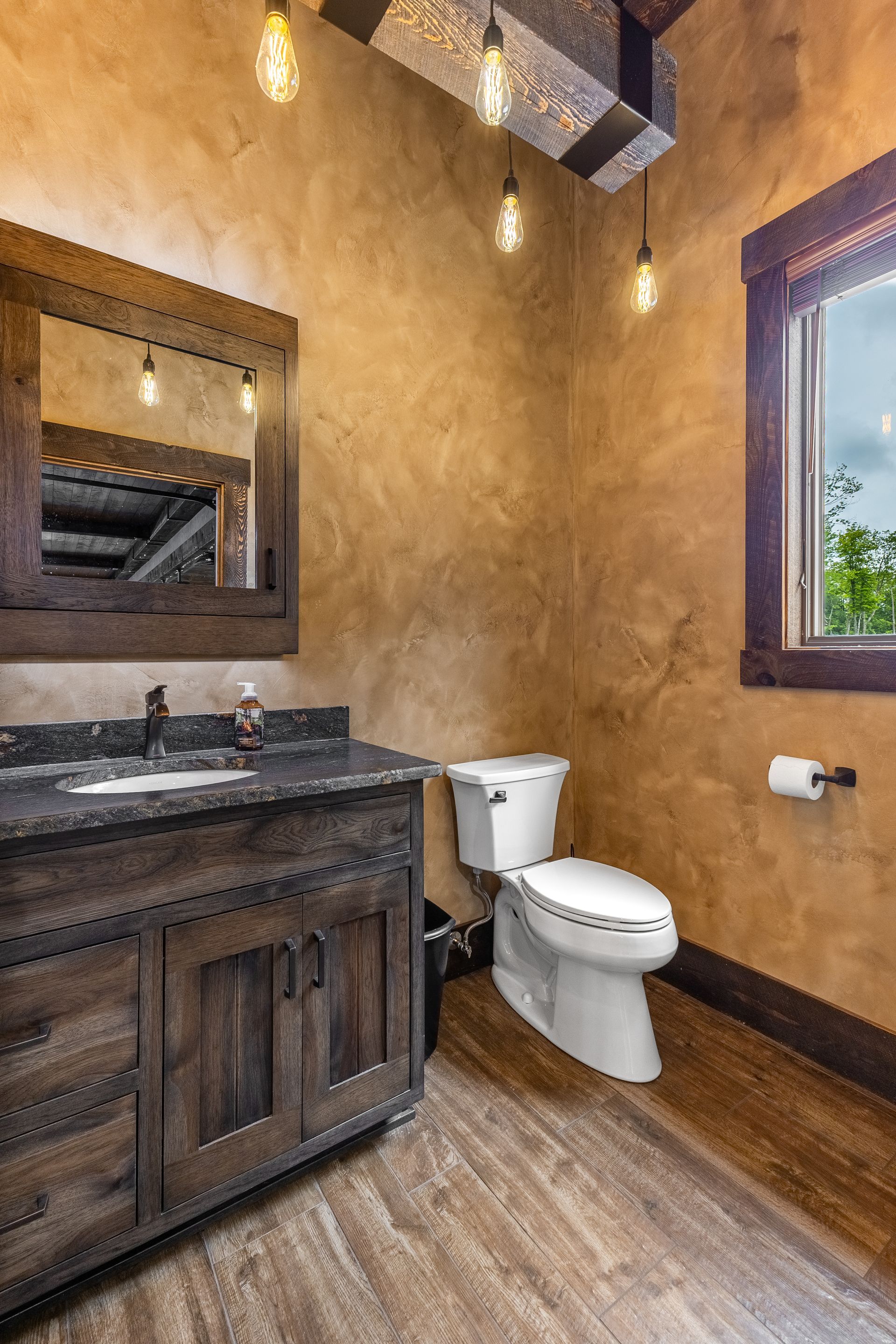 Rustic bathroom with dark wood vanity, white toilet, tan walls, and a window