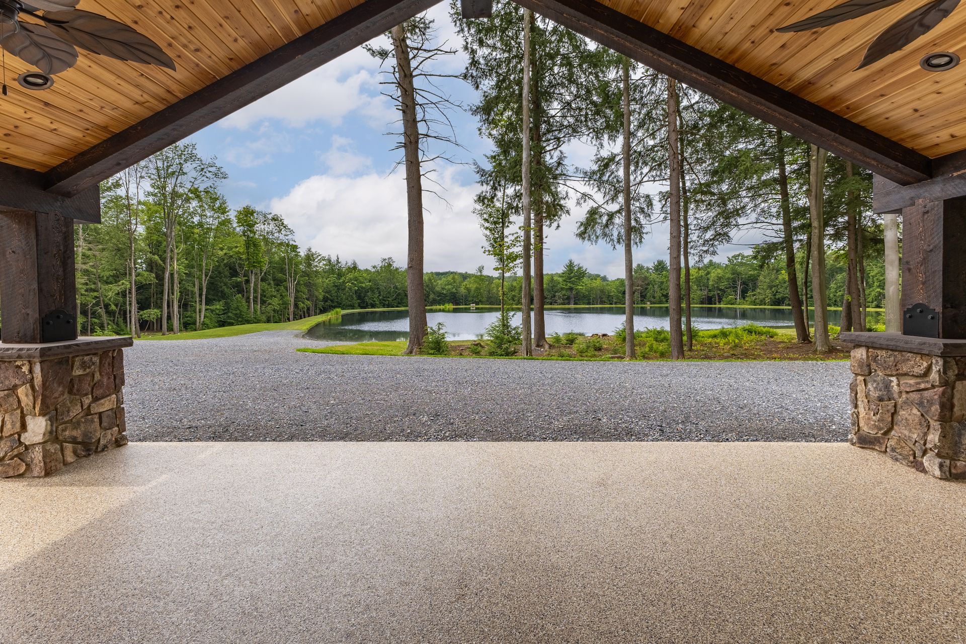 Covered open pavilion overlooking a gravel area, trees, and a lake under a wooden roof