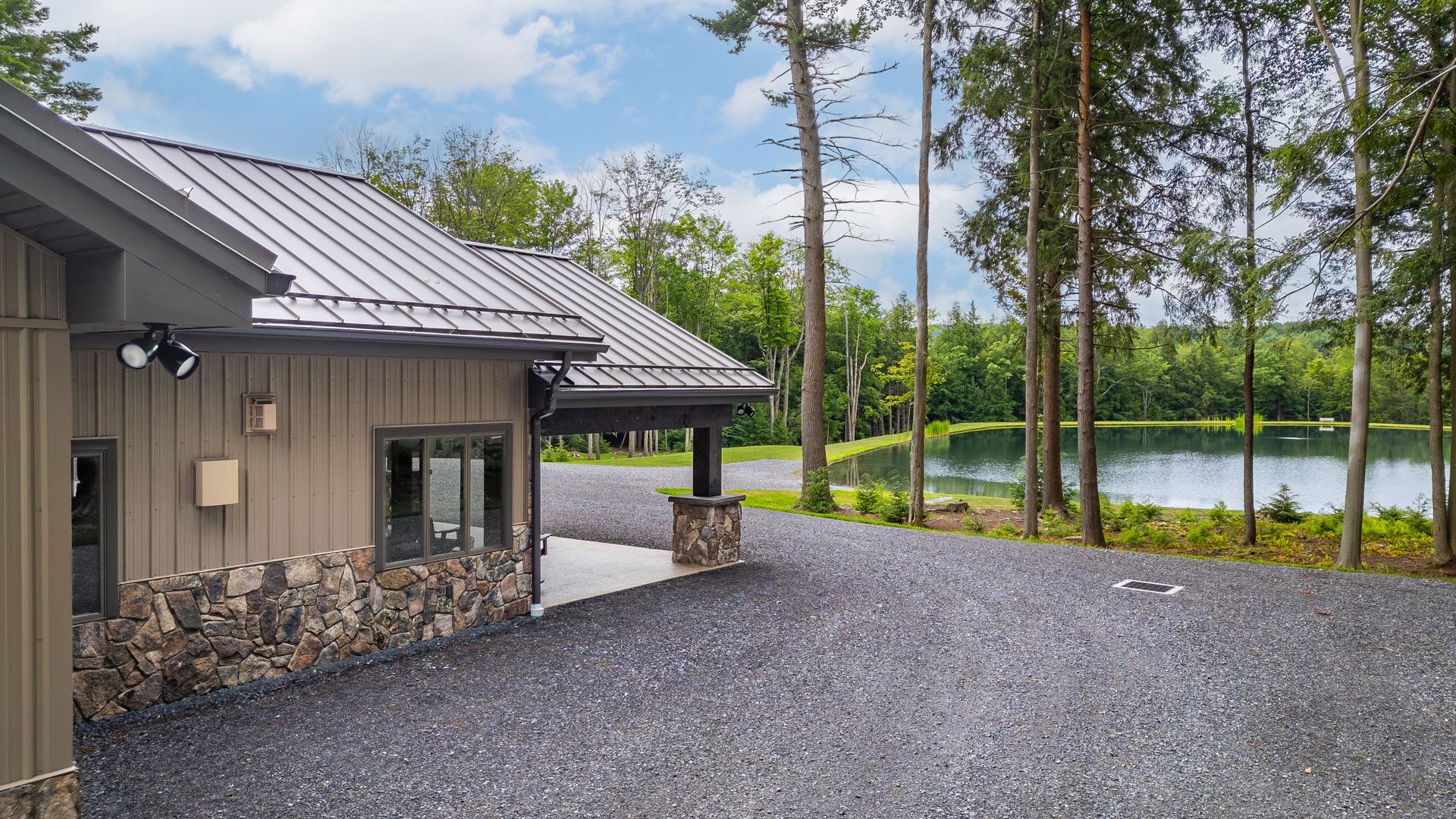 Cabin beside a gravel lot and lake, with trees and cloudy sky in the background