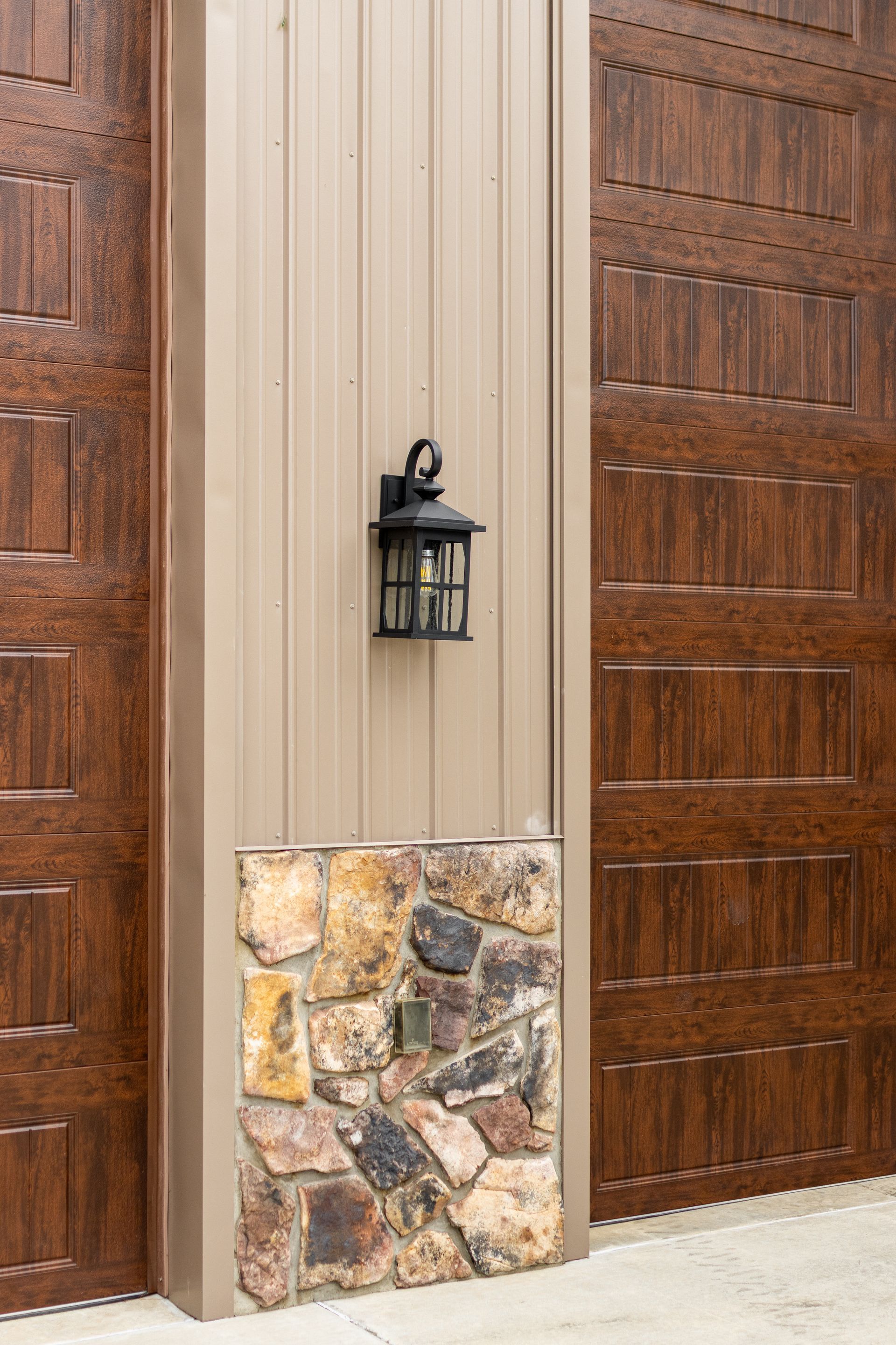 Front entry with tan siding, stone base, and brown paneled garage door beside a black wall lantern