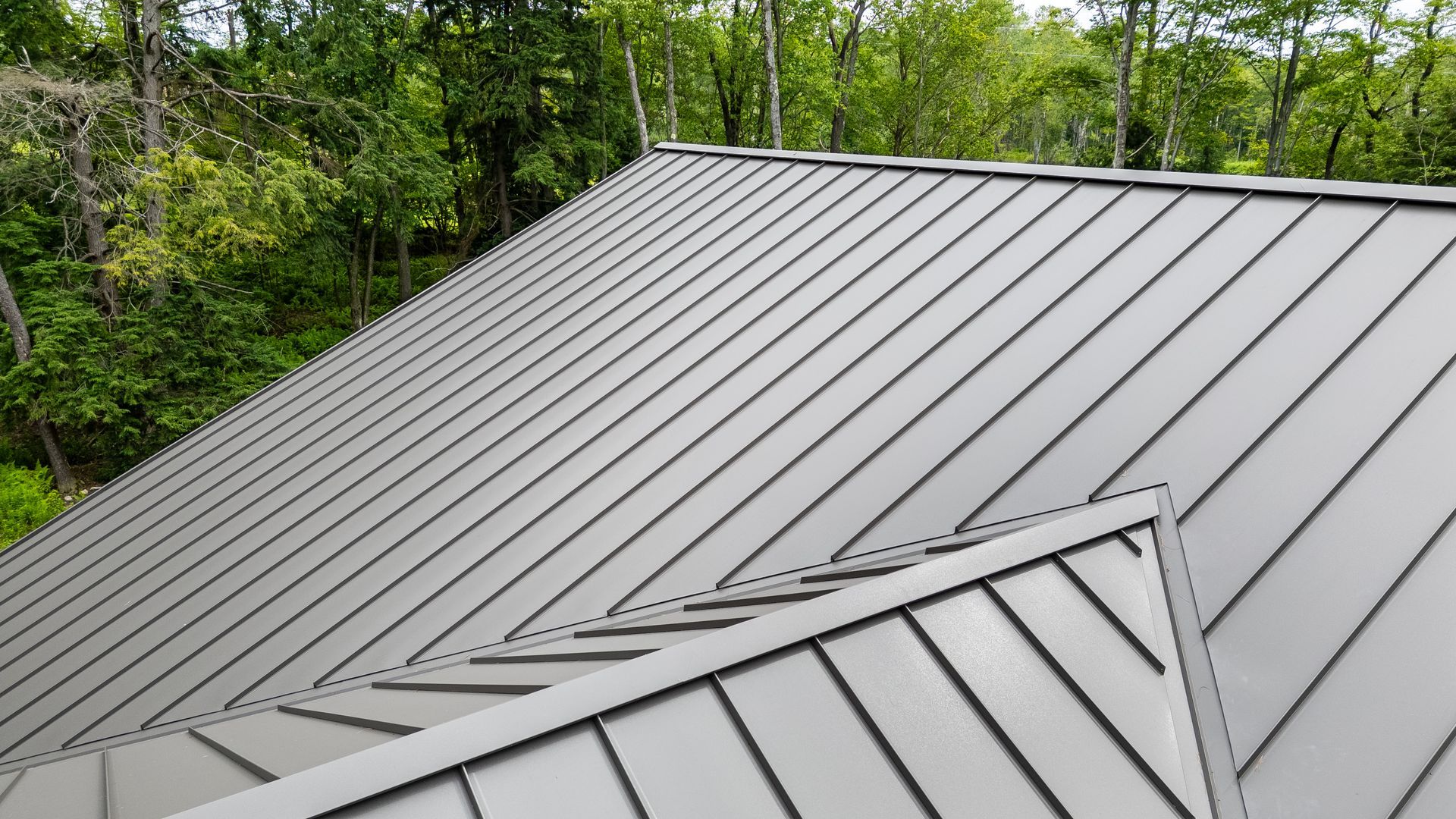 Gray metal roof with a small dormer, backed by green trees under daylight