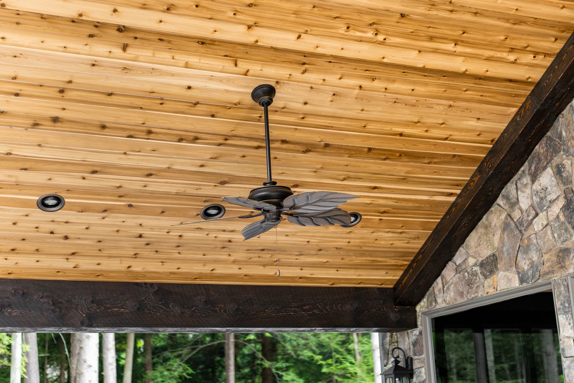 Wooden patio ceiling with a black ceiling fan and dark beams, overlooking green trees.