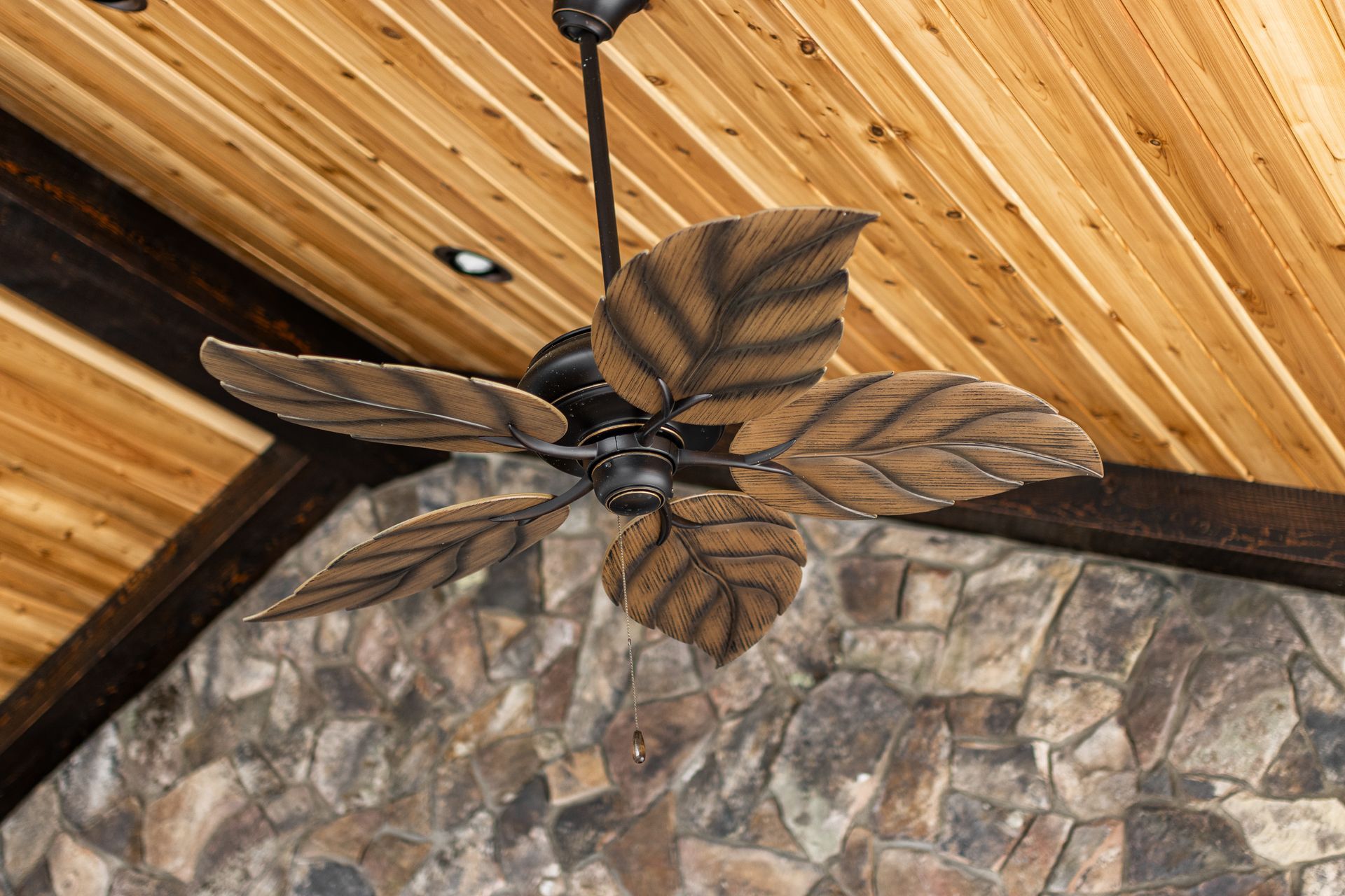 Ceiling fan with dark blades mounted on a wood-planked ceiling above a stone wall.