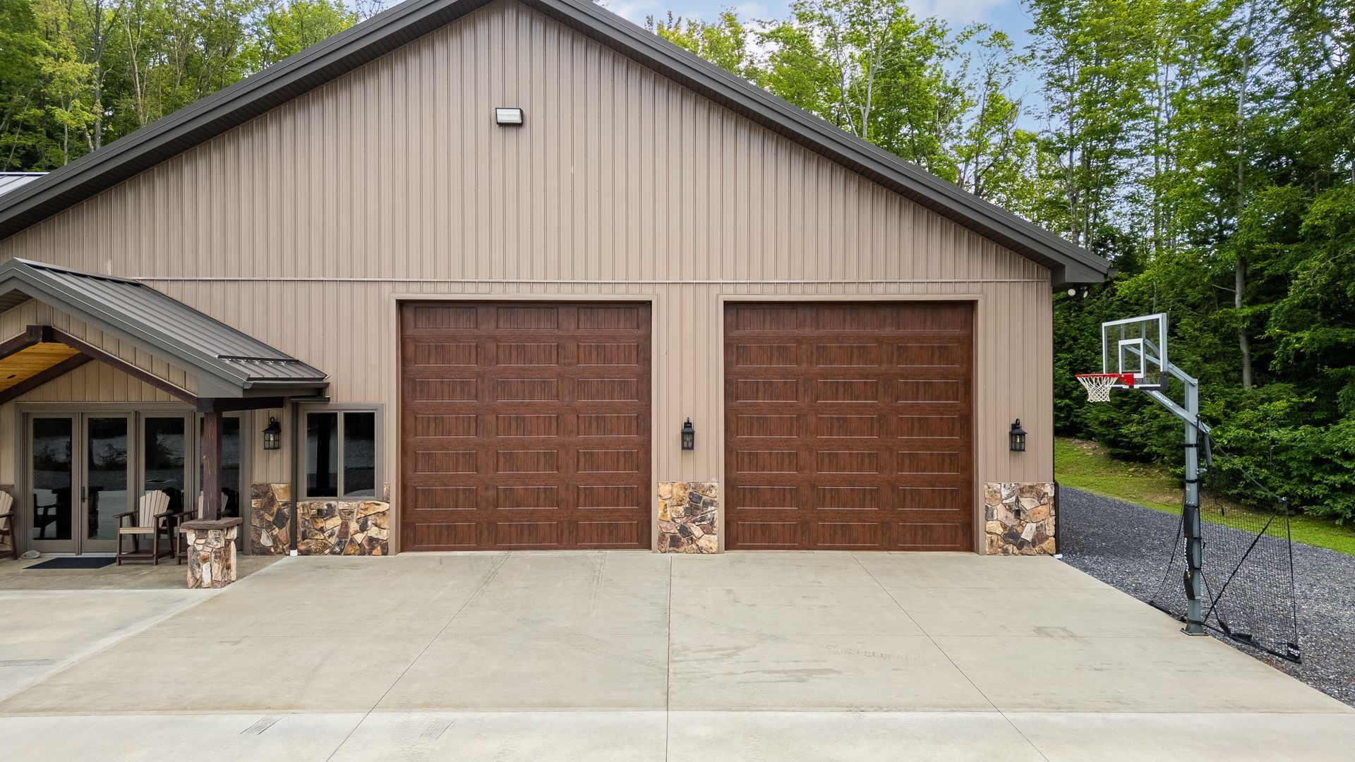 Large beige garage with two brown doors, concrete driveway, and a basketball hoop in front of trees.