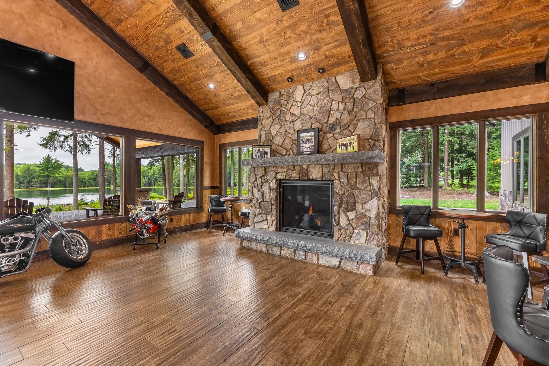 Rustic living room with stone fireplace, wood ceiling, large windows, and hardwood floor