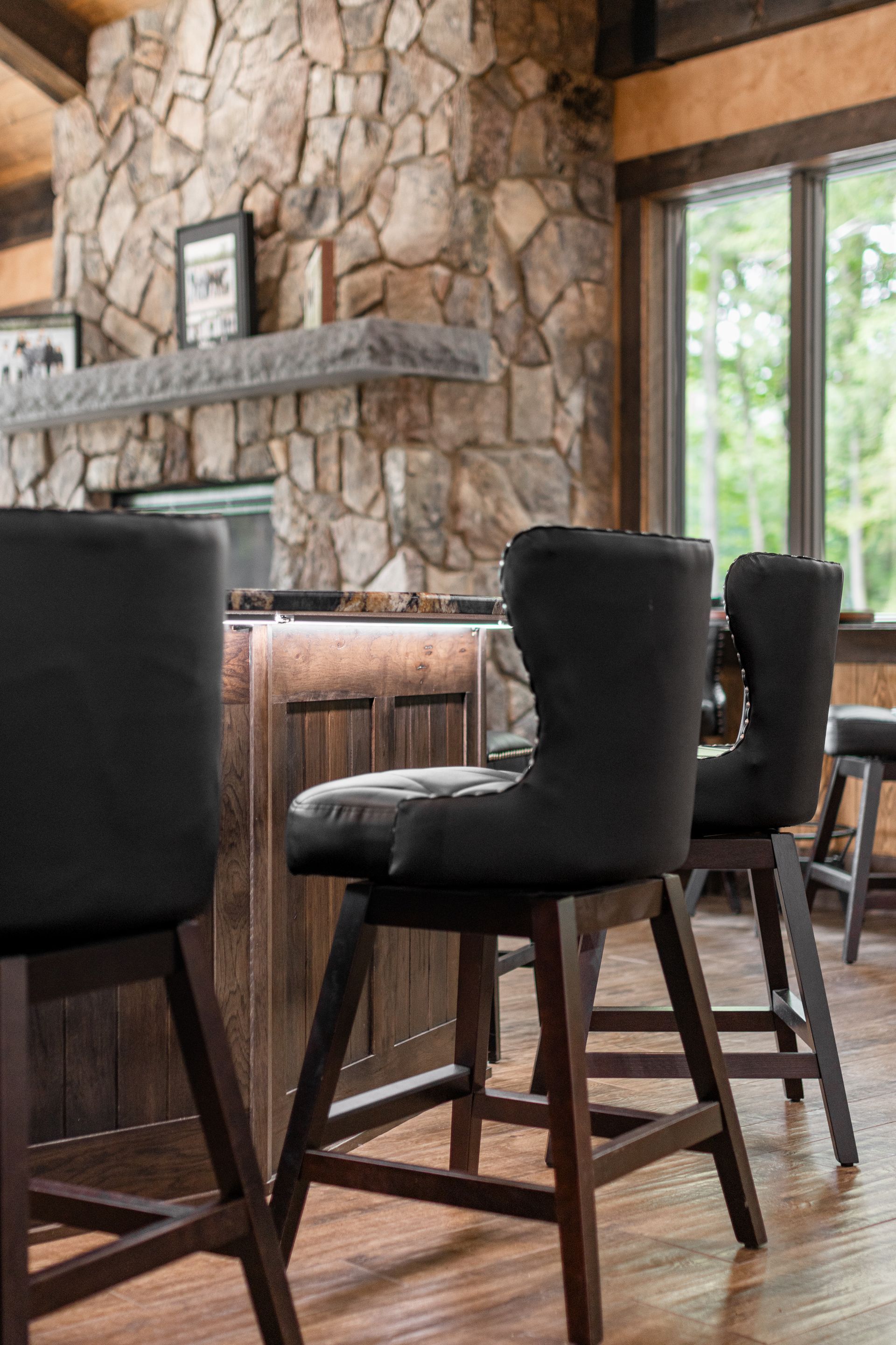 Rustic bar interior with dark stools and a stone fireplace beside large windows