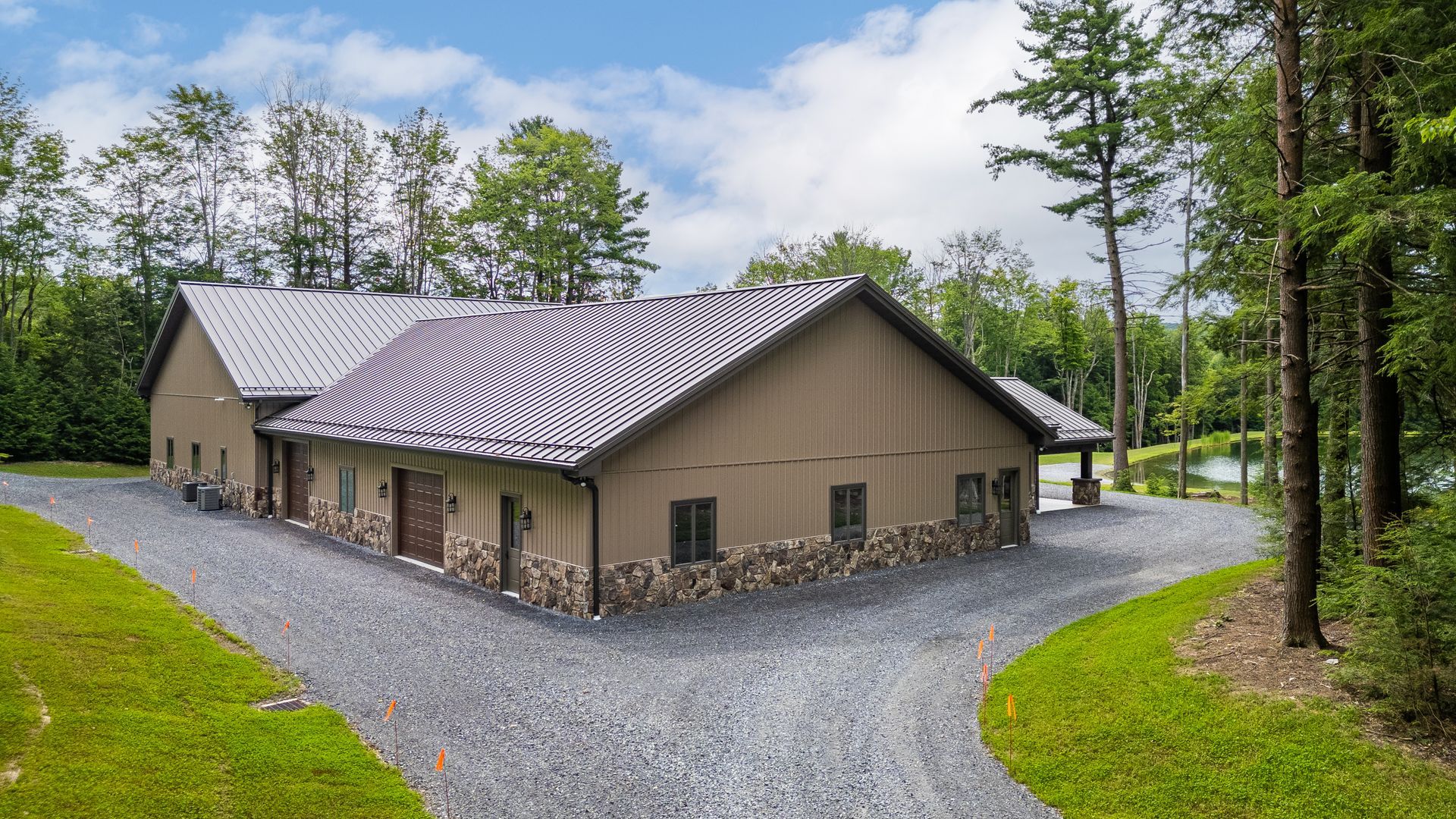 Beige building with a gray metal roof on a gravel drive, surrounded by trees and grass under a blue sky