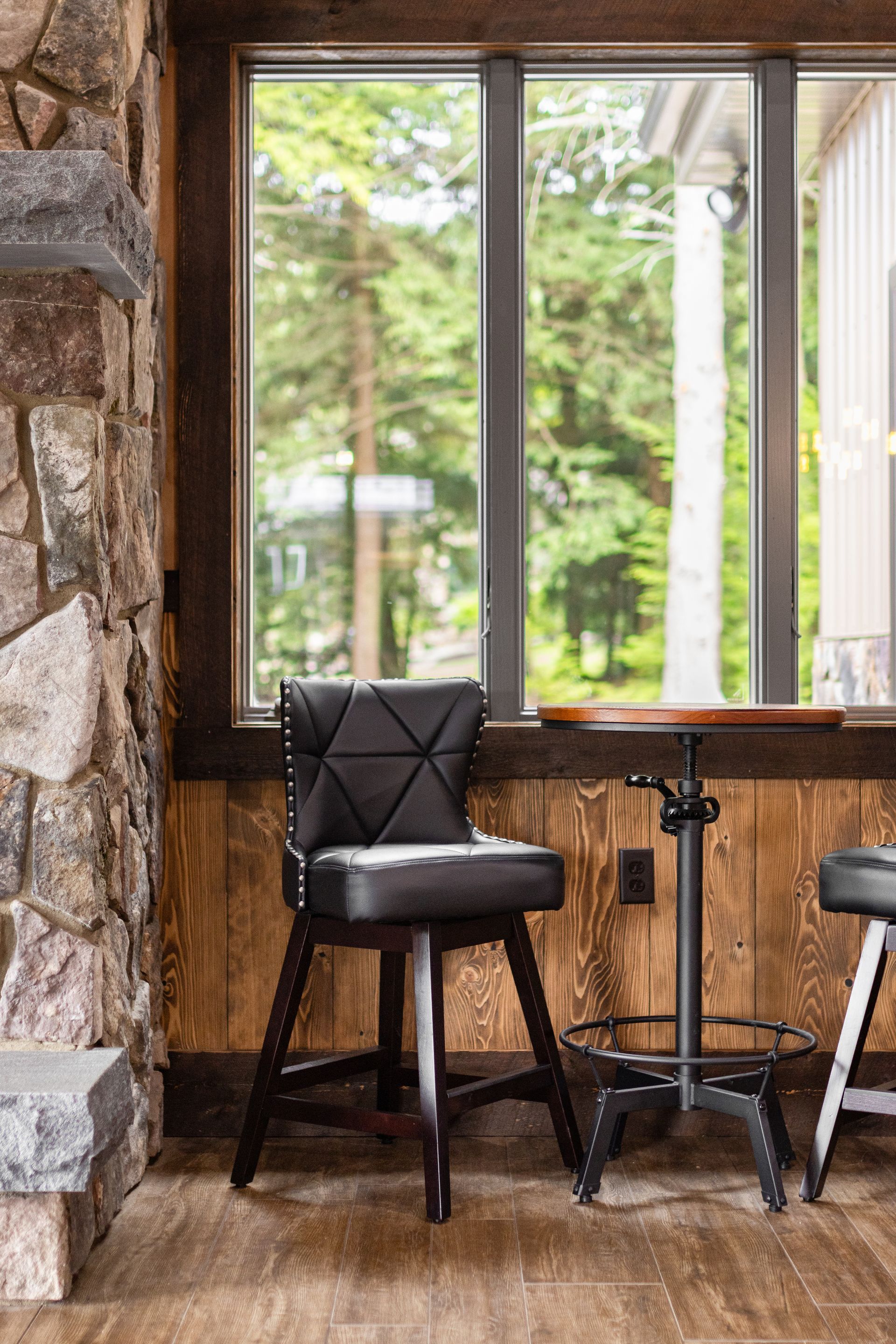 Black bar stool beside a small round table by a large window in a rustic room