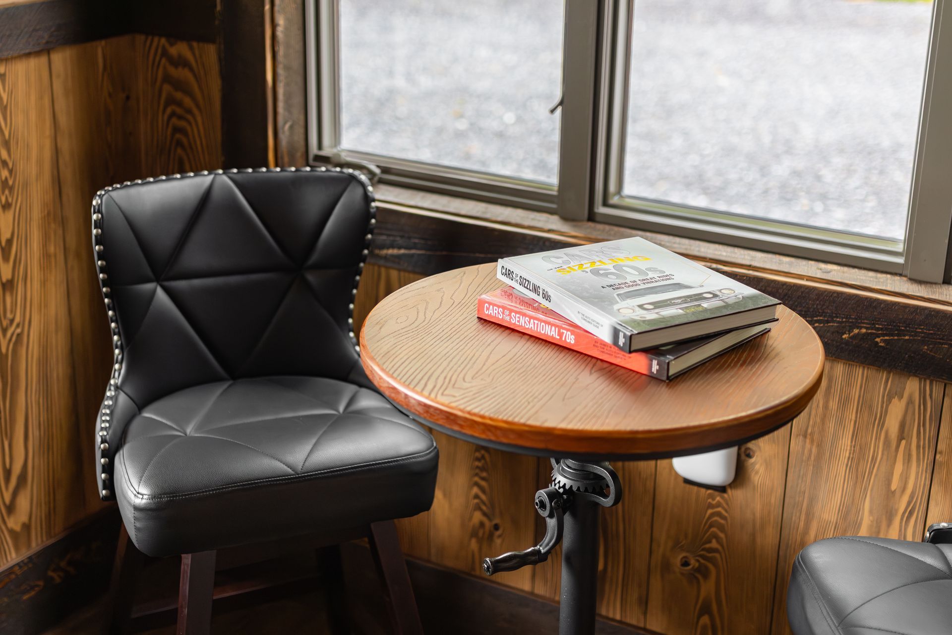 Black chair beside a round wooden table with magazines by a window in a wood-paneled room