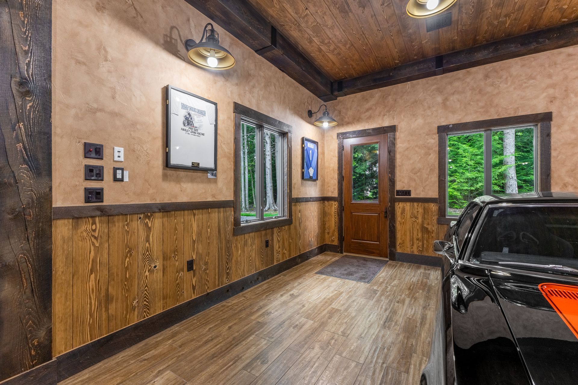 Rustic garage interior with wood-paneled walls, windows, hanging lights, and a black car at right.