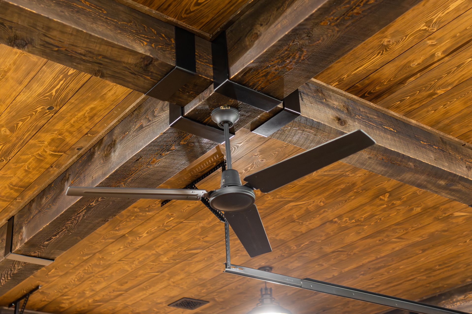 Dark ceiling fan with three blades on a wooden plank ceiling, viewed from below