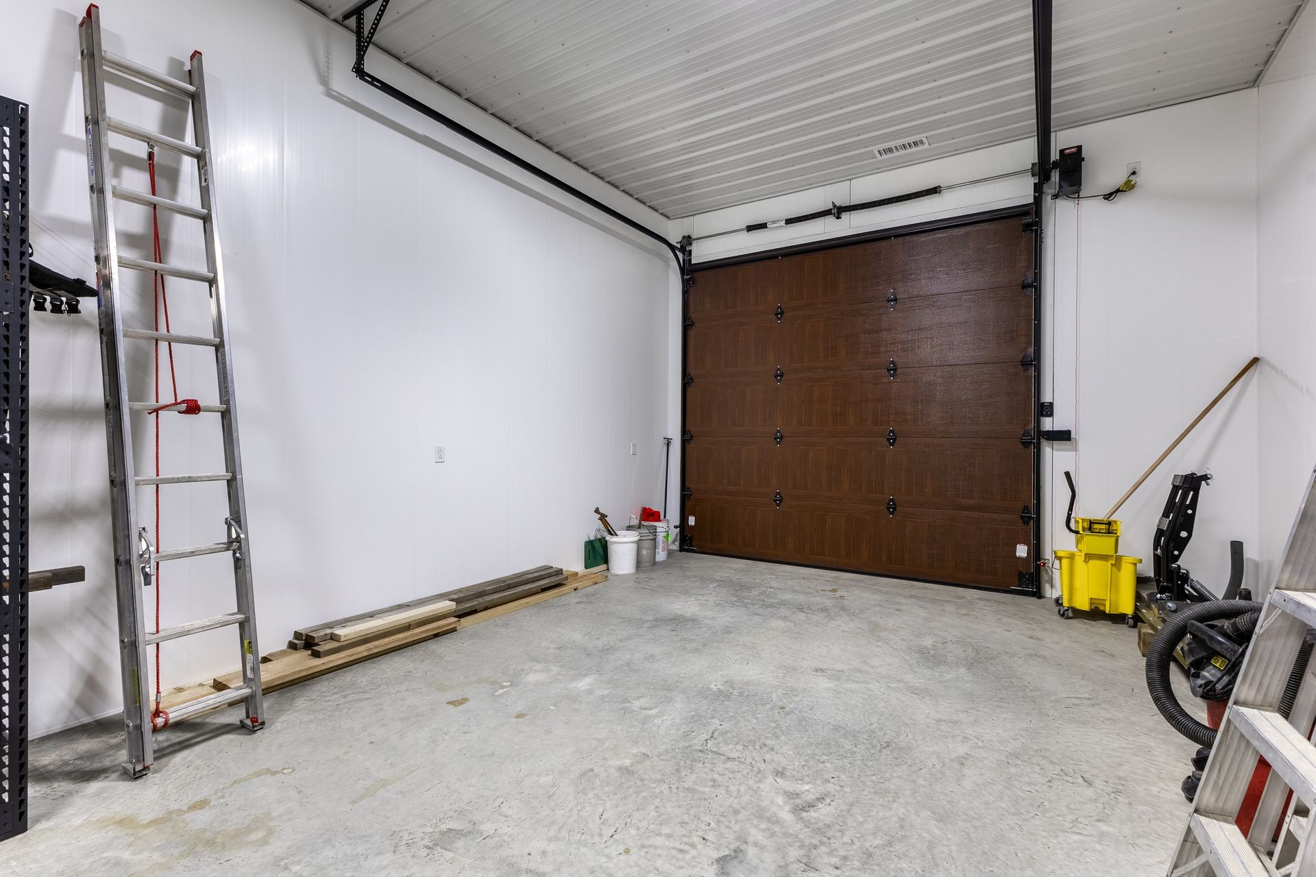 Empty garage with white walls, concrete floor, and a large brown door, with a ladder and tools along the sides