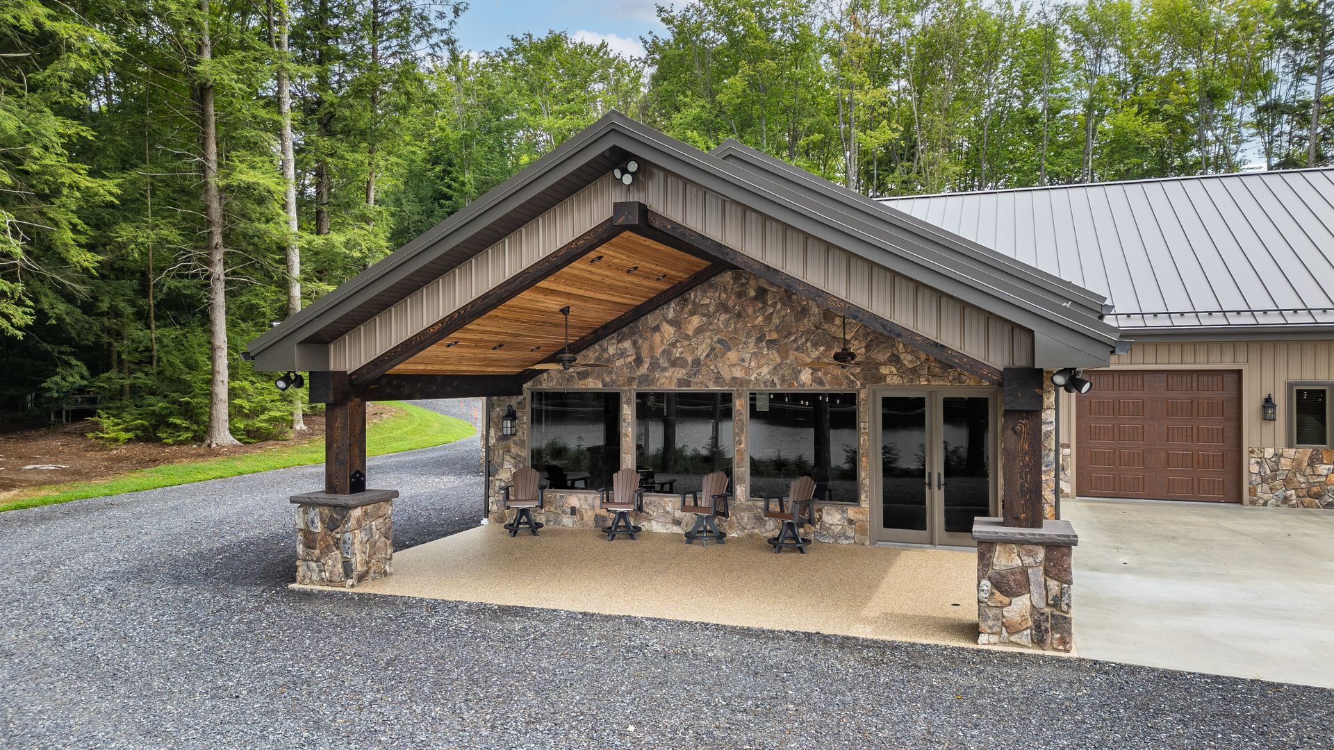Covered lodge-style entrance with stone columns, glass doors, and a metal roof on a gravel driveway.