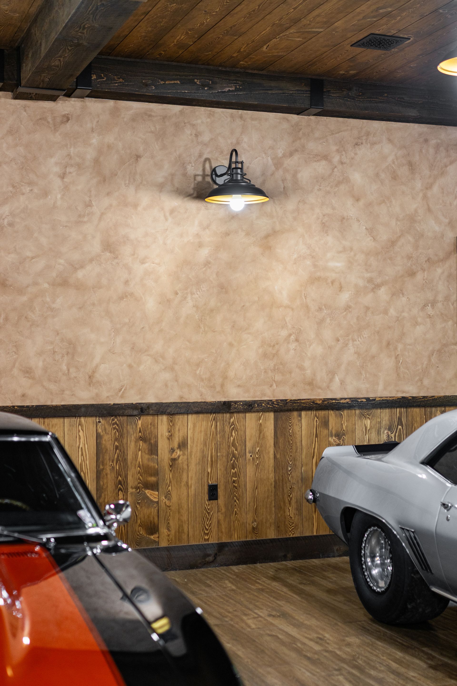 Dim rustic room with tan walls, wood paneling, ceiling lamp, and two classic cars parked inside.