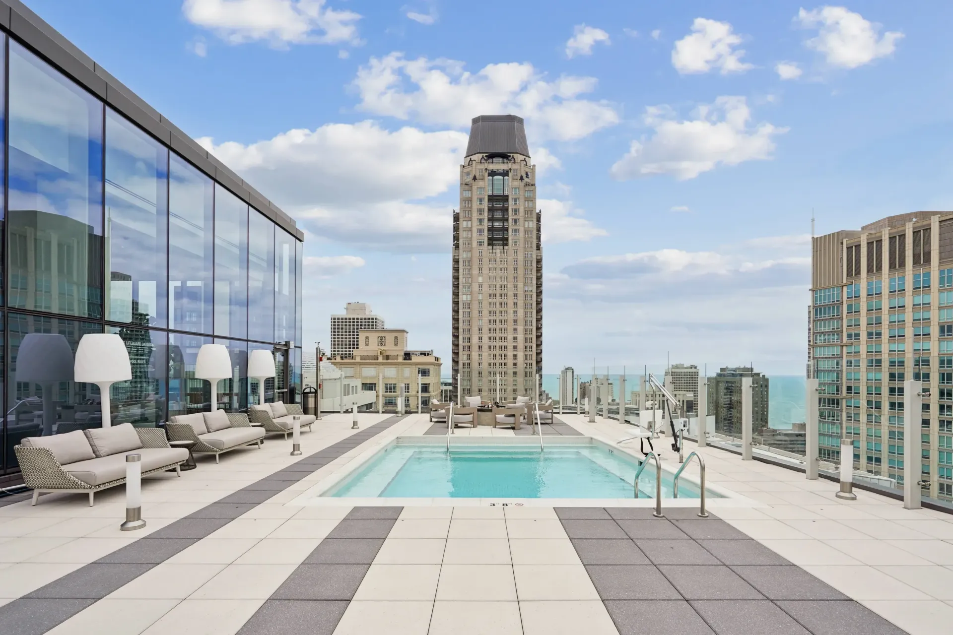Rooftop pool deck with lounge chairs, glass railing, and city skyscrapers under a blue sky