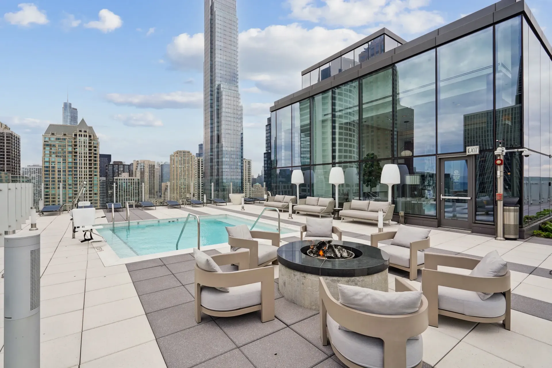 Rooftop terrace with lounge chairs, fire pit, pool, and city skyline beside a glass-walled building