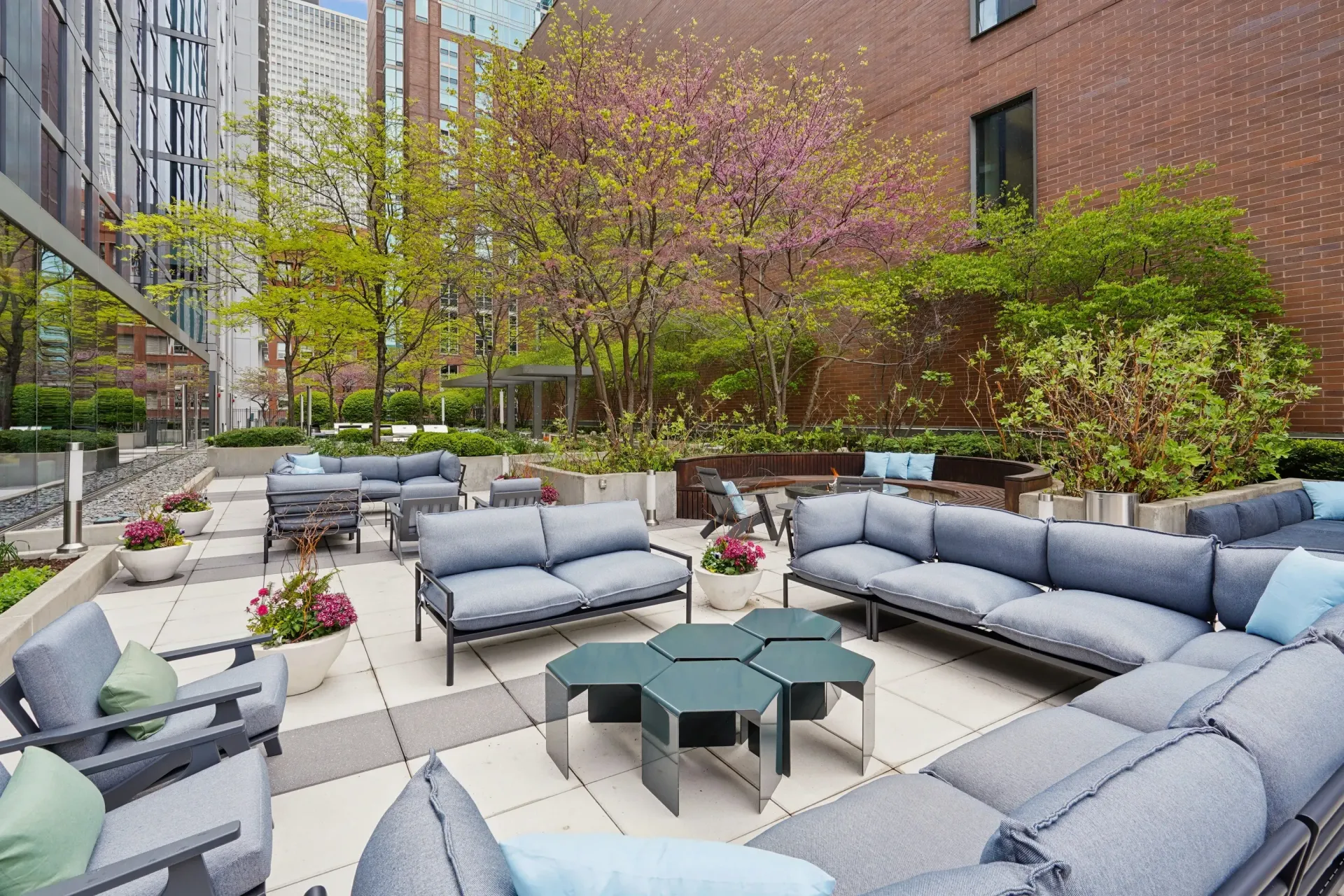 Outdoor apartment courtyard with gray lounge seating, tables, and spring trees beside brick buildings