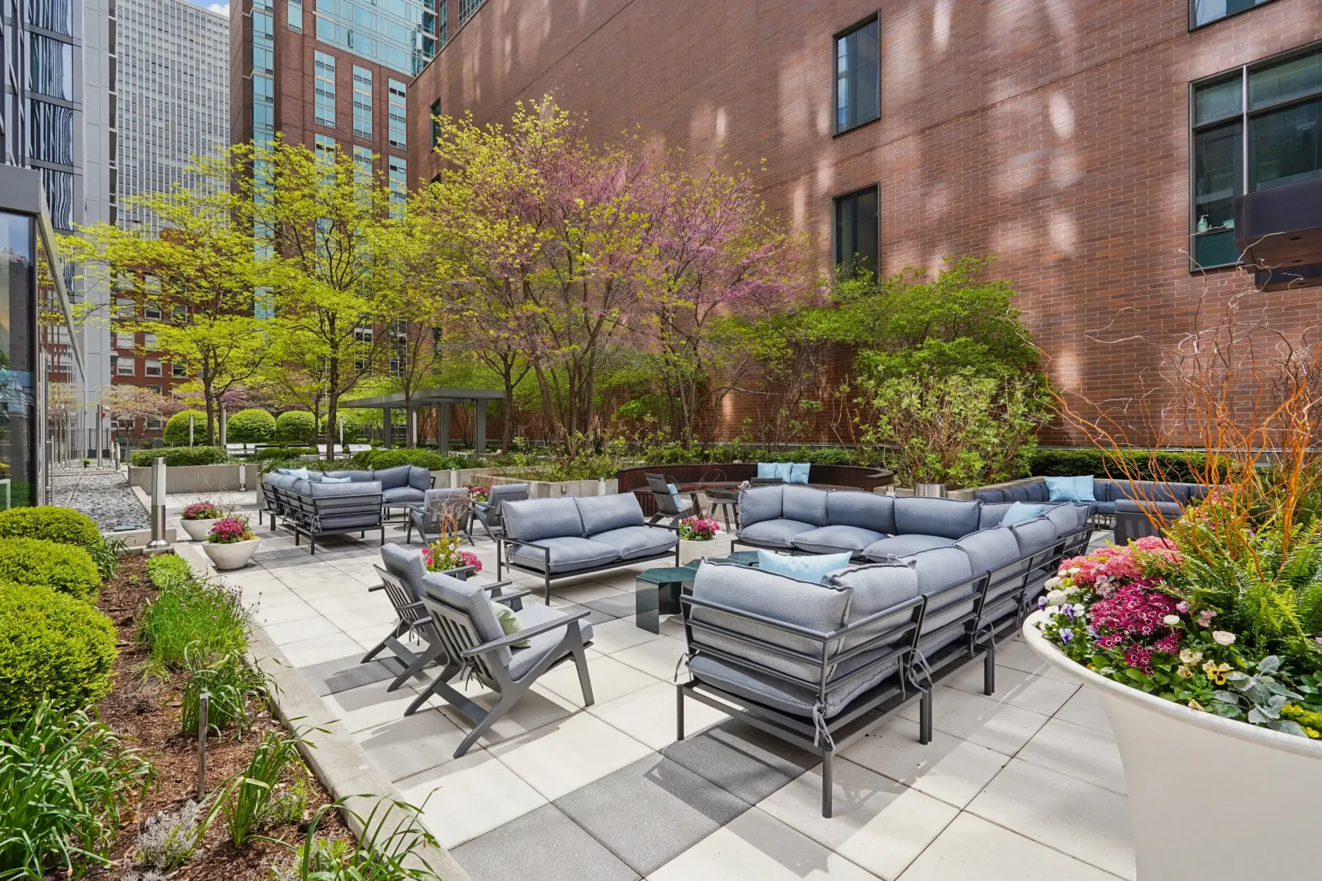Modern outdoor patio with gray seating, planters, and trees beside brick buildings
