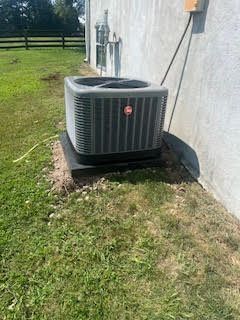 An outdoor air conditioning unit sits on a dark platform next to a beige building. Green grass surrounds it.