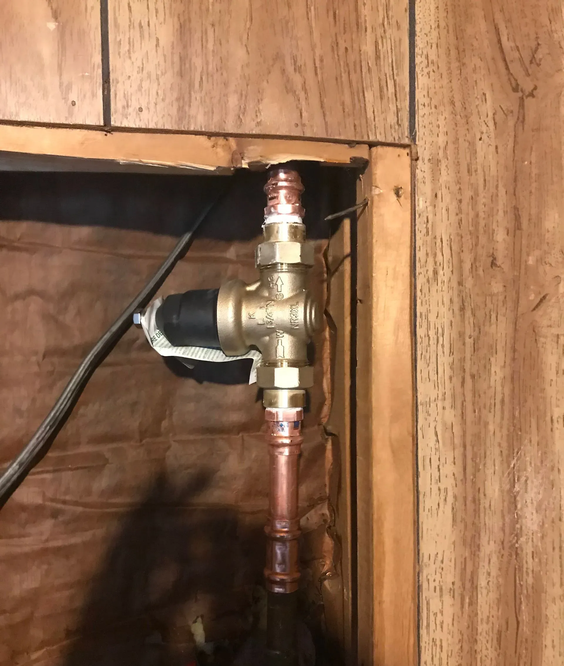 Brass water pressure regulator with copper pipes in a wooden wall cavity.