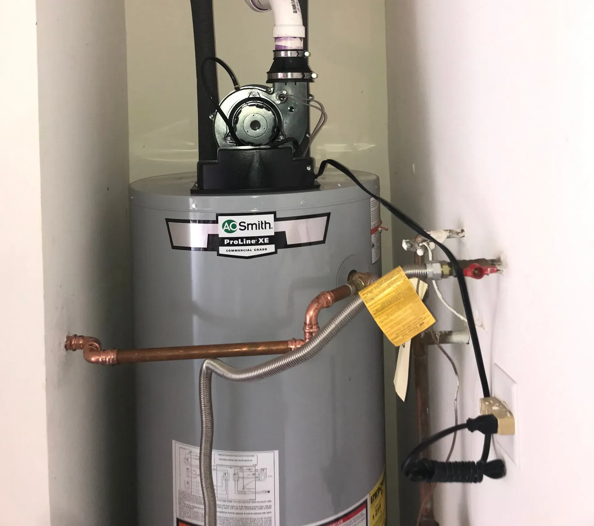 A gas water heater in a white-walled utility closet, with copper piping and a black exhaust pipe.
