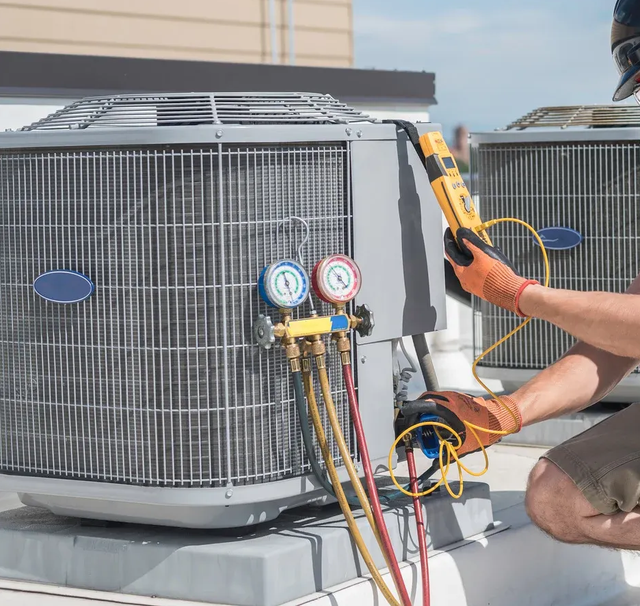 HVAC technician kneels near AC unit on rooftop, using gauges and multimeter, wearing gloves.
