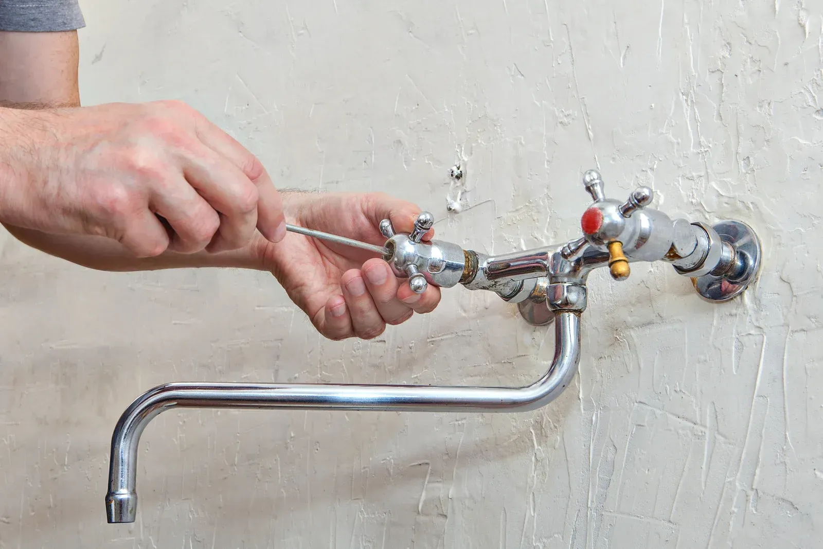 Person using a screwdriver to repair a chrome faucet on a textured wall.