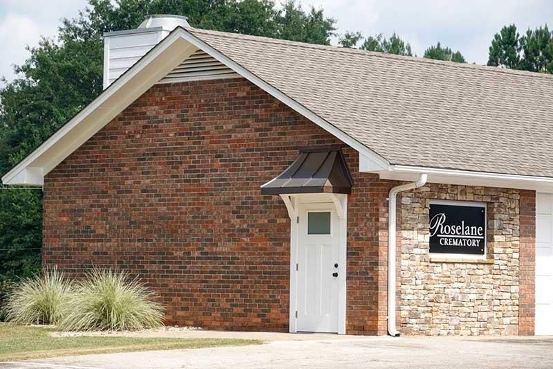 Brick church building with a white door and stone sign, seen from the front under a shingled roof.