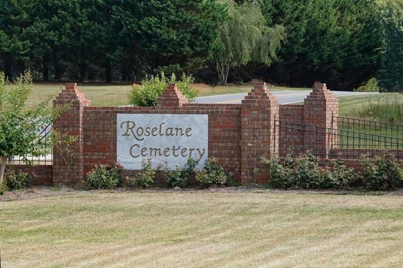 Roselane Cemetery sign on a brick entrance wall beside a grassy field and trees