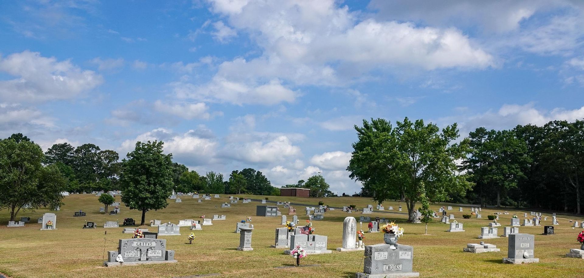 Grassy cemetery with rows of white headstones and trees under a partly cloudy blue sky