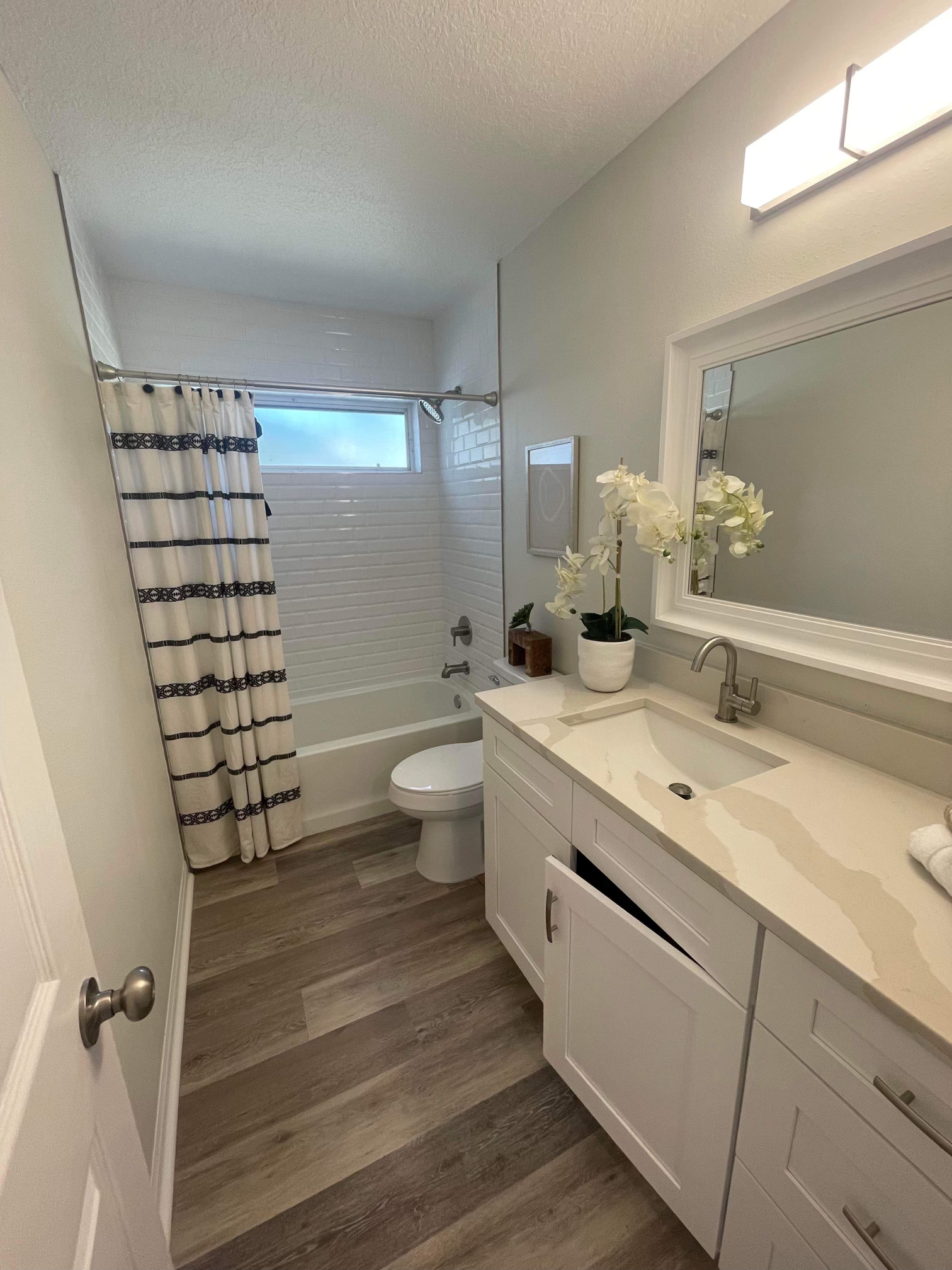 Bathroom with white cabinets, beige countertop, gray wood-look floor, and a tub/shower with striped curtain.