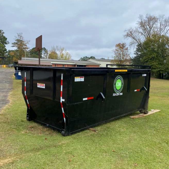 Black dumpster with orange reflectors on grass, outdoors near a court fence and trees.