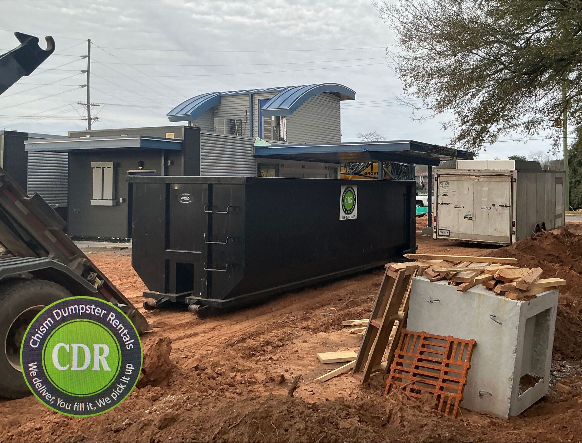 Black dumpster on muddy construction site beside damaged concrete and a partially built house