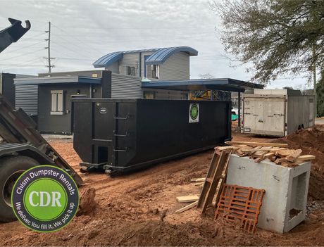 Black dumpster on muddy construction site beside damaged concrete and a partially built house