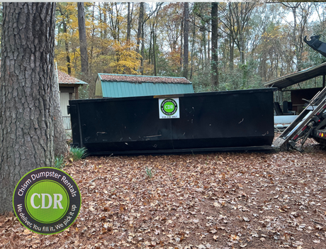 Black dumpster in wooded yard with fallen leaves, CDR logo on side