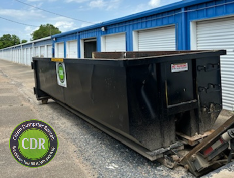 Large black dumpster beside blue storage units on a gravel lot