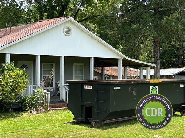 White house with porch beside a large green dumpster in a grassy yard