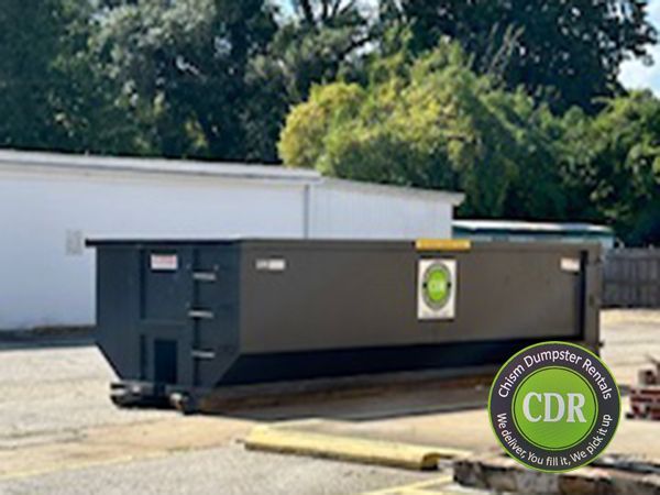Large black dumpster in a parking lot beside a white building, with trees in the background.
