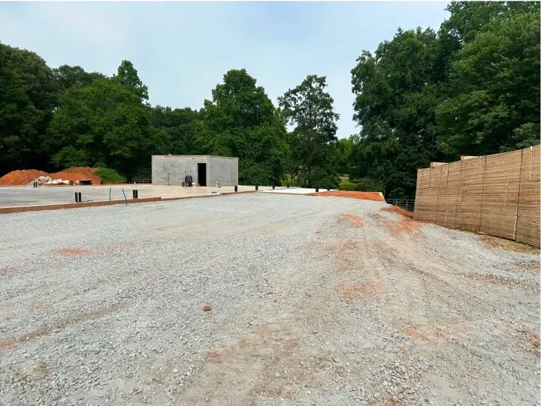 Gravel parking area with a small concrete building and trees in the background under a cloudy sky.