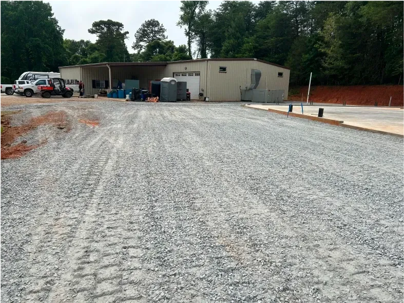 Gravel parking area in front of a tan building with several vehicles parked to the left, trees in background.