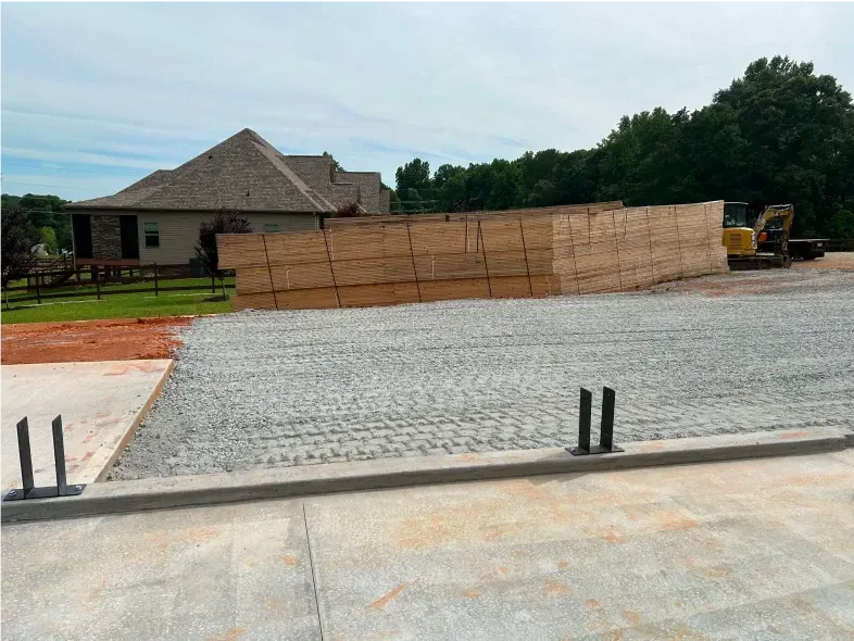 Construction site with a stack of lumber, a gravel base, and a house in the background.