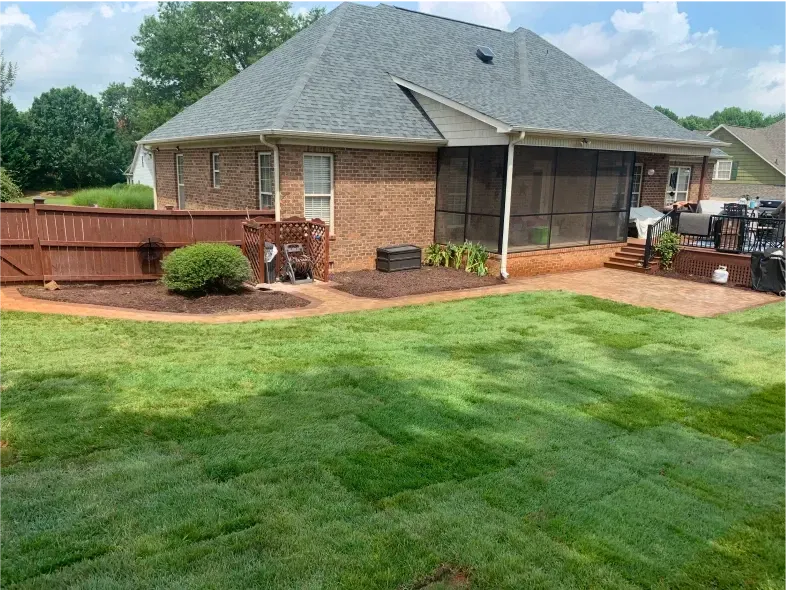 Backyard with brick house, green lawn, brown fence, and screened porch on a sunny day.