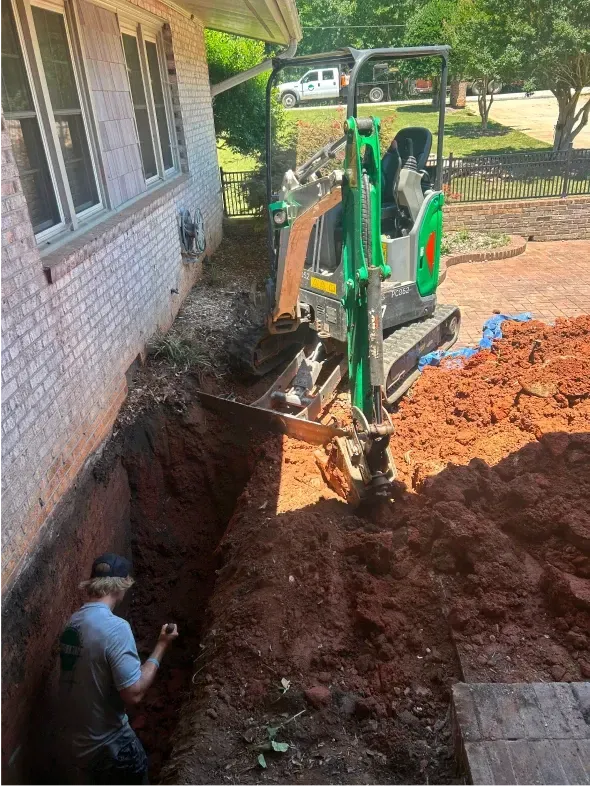 A mini-excavator digs beside a brick building. A person is in the trench, observing. Sunny outdoor setting with dirt.