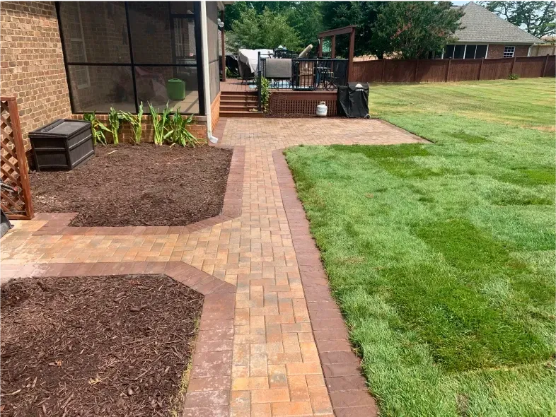 Brick patio with path and planted beds next to green lawn, leading to a wooden deck.