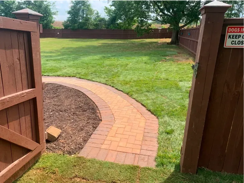 Brown brick path curves through a grassy yard, enclosed by a brown wooden fence with an open gate.