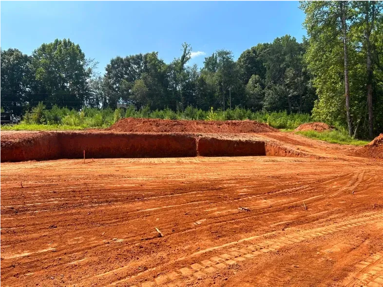 Cleared red dirt construction site with a berm, trees in the background, blue sky.