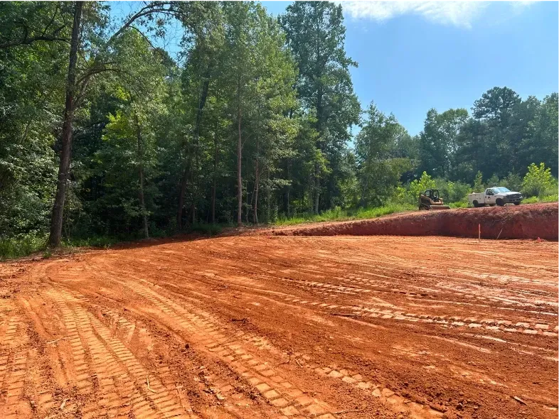 Cleared red dirt lot with tire tracks, trees in the background, clear sky.