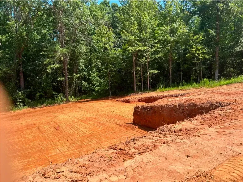 Cleared land for construction, with orange soil and a backdrop of green trees.