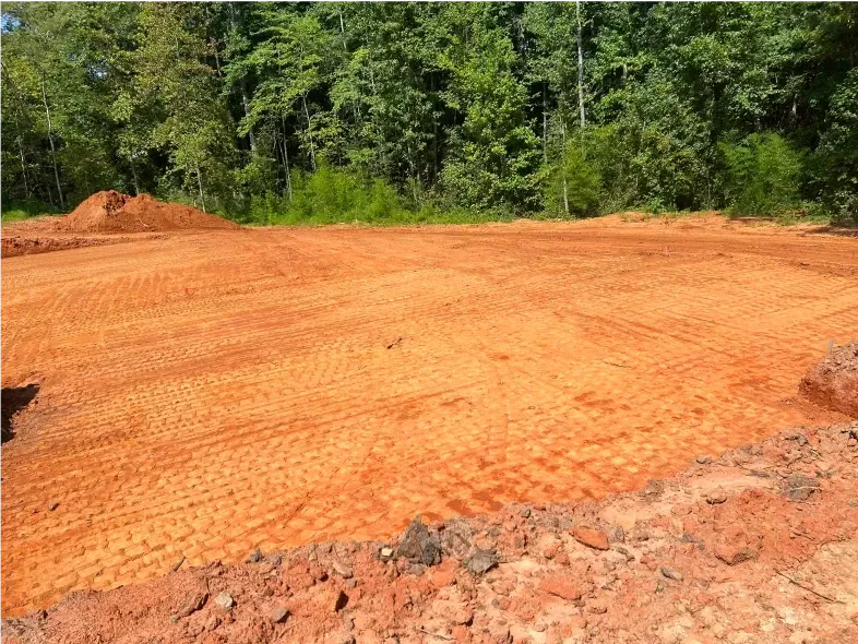 Cleared red dirt lot with tire tracks, trees in the background.